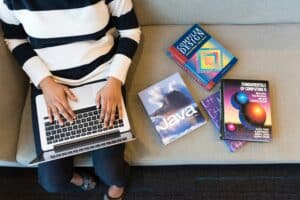 A person in a striped sweater sits on a sofa using a laptop, with several programming books nearby.